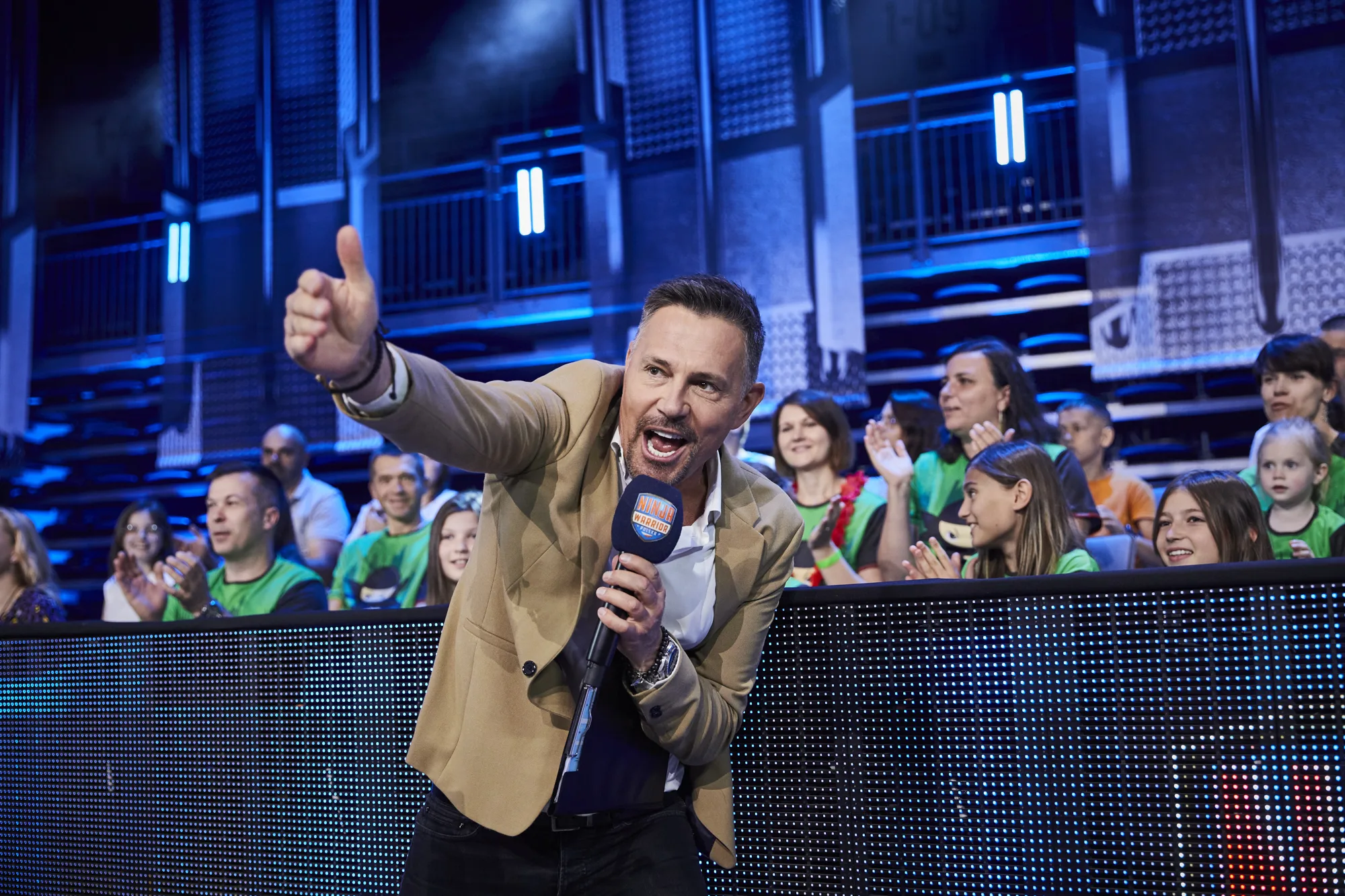 Prowadzący z mikrofonem Ninja Warrior na tle rozentuzjazmowanej publiczności — dzieci z Ninja Club Kraków